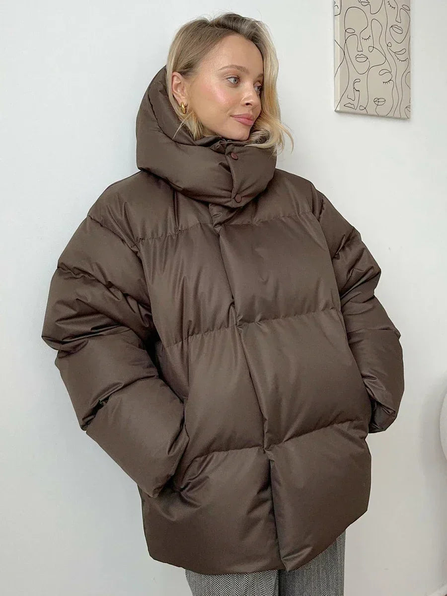Vrouw draagt een stijlvolle gewatteerde winterjas, ideaal voor koude dagen. De jas is oversized en comfortabel, met een royale capuchon voor extra warmte en bescherming tegen de kou. Ze combineert de jas met een gebreide muts, zonnebril en casual outfit, wat een modieuze en warme uitstraling creëert. Perfect voor winterse wandelingen en dagelijkse activiteiten in de kou.
