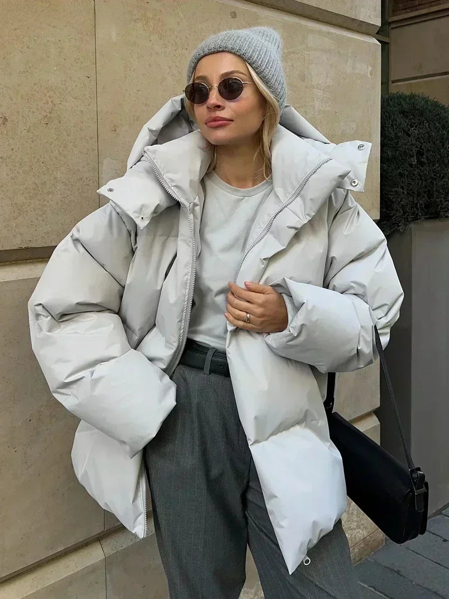 Vrouw draagt een stijlvolle gewatteerde winterjas, ideaal voor koude dagen. De jas is oversized en comfortabel, met een royale capuchon voor extra warmte en bescherming tegen de kou. Ze combineert de jas met een gebreide muts, zonnebril en casual outfit, wat een modieuze en warme uitstraling creëert. Perfect voor winterse wandelingen en dagelijkse activiteiten in de kou.